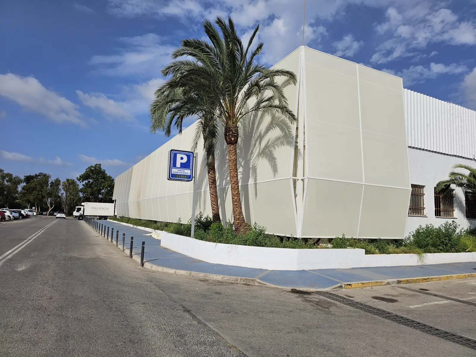 Fachada textil blanca junto a un aparcamiento con palmeras
